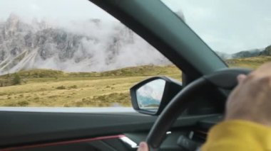 Man drives car on highway against giant Italian Alps surrounded by fog. Hands of driver holding steering wheel and scenic view outside window closeup