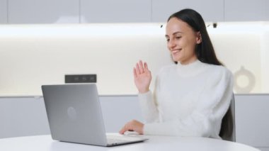 Young female team leader greets participants of webinar gladly sitting in light room. Smiling woman happy to share knowledge. Online communication