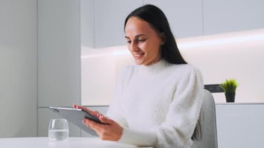 Young brunette woman smiles gladly chatting with friends on social networks via tablet. Online communication using modern gadget close side view