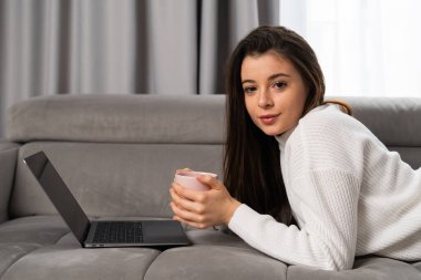 Portrait of a beautiful young girl lying on a sofa with a cup of tea and a laptop. 