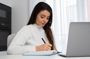 Intelligent female student writing notes to textbook sitting at home and preparing to the exam. 