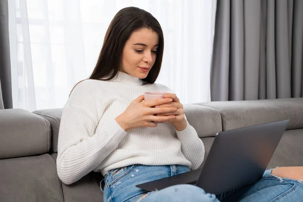 Young woman relaxing on sofa holding a cup. Woman watching on the screen of laptop.
