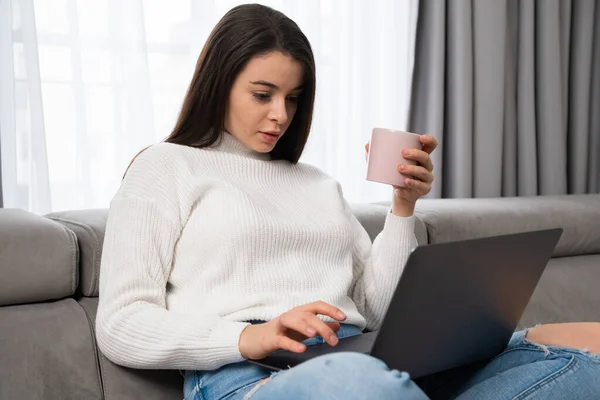 Cheerful woman with cup of tea using laptop in living room.