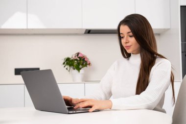 Cheerful brunette using a laptop while sitting in the kitchen. Young woman chatting with her friends via social networks. 