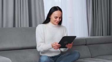 Young woman communicates with friends on social networks sitting at home. Lady works on modern tablet computer smiling contentedly