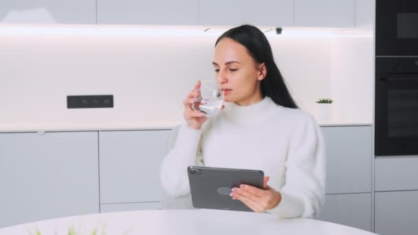 若い女性はタブレットコンピュータで働くガラスから水を飲む 成功した女性はテーブルに座ってビジネス上の問題を解決することを考える — ストック動画