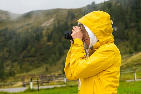 Woman in the hood and yellow sport jacket makes photos of meadows of Alps. Young woman tourist captures moments with camera and explores foggy hillside in mountains