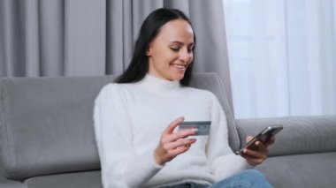 A woman purchases gifts via the Internet using a mobile phone and credit card sitting in the living room for Happy Valentines Day. 