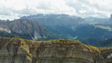Green valley and large forests behind rocky Seceda ridgeline covered with light haze. Scenic Italian Alps under cloudy sky aerial view