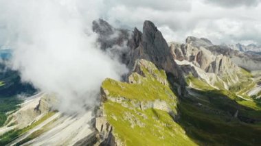 Dense foggy cloud descends on giant Seceda ridgeline with green slopes and bare peak. Cloudy sky above touristic Italian Alps aerial view