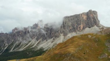 High rocky cliffs of Giau pass in Dolomites surrounded by light clouds in sky. Mountain valley with trees and meadows surrounding hill slopes