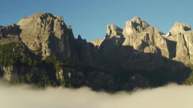Rocky peak of giant Alps against clear blue sky at sunrise. Dense fog surrounds high mountains on sunny summer morning aerial view