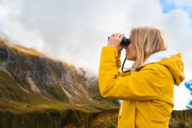 Young blonde woman hiking in mountains in the Giau Pass and looking through binoculars. Wanderlust concept. 