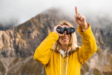 Smiling girl looking through binoculars and showing by finger in the distance with amazing mountains covered by clouds on the background. 