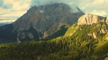 Giant forestry mountains with bare rocky peaks under clouds at sunset. Evergreen coniferous trees grow in forests in Italian Alps aerial view