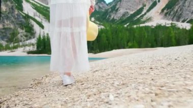 Close up of womans legs walking along a mountain lake in a white dress holding a straw hat in her hand. Travel concept. 
