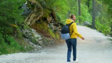 Kadın turist, yeşil ağaçlarla ormanda yolda yürüyor. Sırt çantalı bir kadın göl manzarası boyunca yabanda yürüyüş yapmaktan hoşlanıyor.