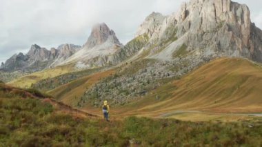 Turist patikada duruyor ve güneş ışığıyla aydınlatılan yüksek kayalık kayalıkları keşfediyor. Sırt çantalı gezgin dağlara bakarak sonbahar otlaklarında yürüyor.