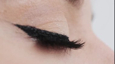 Uzun yapay kirpikleri ve siyah oku olan genç bir kadının gözü. Kahverengi gözlü bir kadın beyaz arka planda göz kırpıyor.
