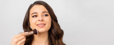 Yüzüne yakın makyaj fırçası tutan mükemmel ciltli genç bir kadın. Gri arka planda fotokopi alanı olan.