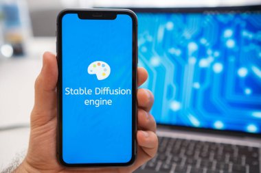 Bir adam ekranda sabit difüzyon motoru logosu olan bir akıllı telefonu tutarken, arka planda bir dizüstü bilgisayar sıçrama ekranında bir nöron görüntülüyor. Mart 2023, Prag, Çek Cumhuriyeti 