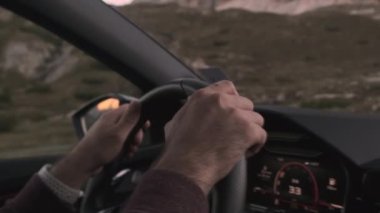Bir adam arabasını dağ yolunda sürüyor, elleri ustalıkla direksiyonu çeviriyor bulutsuz bir gökyüzünün altında çarpıcı bir dağ sırasını geçerken..