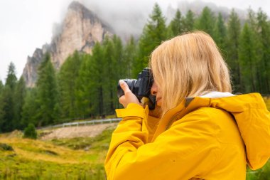 Blonde lady makes photos of meadows of Alps. Young woman tourist captures moments with camera and explores foggy hillside in mountains