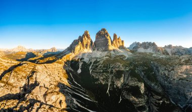 Güneş Tre Cime di Lavaredo 'nun görkemli dağlarının üzerinden yükseliyor. Güneşin doğuşunda açık gökyüzünün altındaki yüksek kayalık tepeler arkadaki hava manzarası aydınlatıyor.