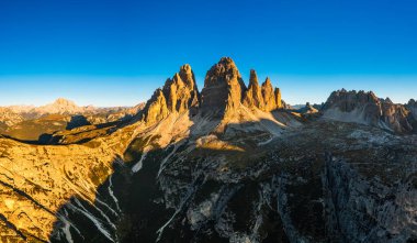 Çıplak kayalık dağların devasa sivri tepeleri mavi bulutsuz gökyüzünün altında sıralanır. Gün doğumunda Tre Cime di Lavaredo 'nun manzarası.