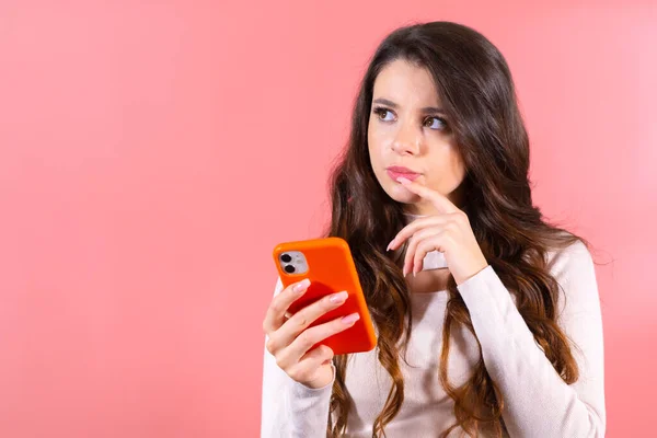 stock image Portrait of thoughtful attractive young brunette woman with a smartphone, chatting with friends looking up empty space or thinking about new post isolated on pink color background. 