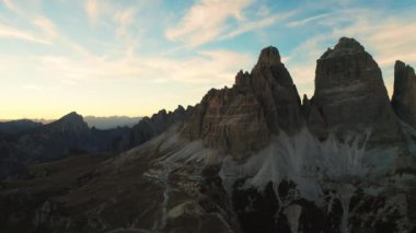 Canlı günbatımı renkleri kayalık dağ sıralarını boyar. Tre Cime di Lavaredo eteklerinin siluetli kumla kaplı yamaçları arka plandaki aydınlık hava manzarasında göze çarpıyor..