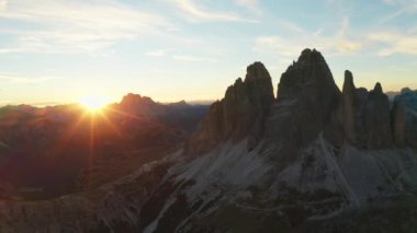 Güneş batıyor, kayalık dağları aydınlatıyor. Tre Cime di Lavaredo eteklerinin siluetli kumla kaplı yamaçları arka plandaki hava manzarasında büyülenir..