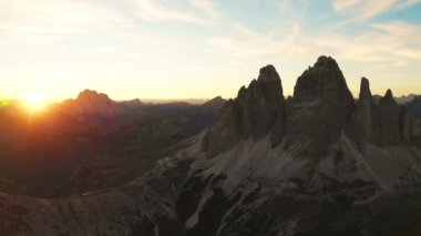 Gün batımı, kayalık dağ sıralarında sıcak bir parıltı yayar. Tre Cime di Lavaredo eteklerinin siluetli kumla kaplı yamaçları arka planda aydınlık hava manzaralı.
