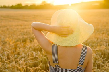 Hasır şapkalı bir kadın buğday tarlasında günbatımının tadını çıkarıyor..