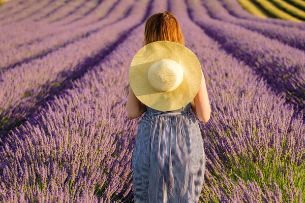 A brunette woman in a sundress and a straw hat stands in a lavender field.