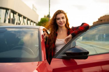 Canlı kızıl saçlarıyla genç bir kadın, sokakta parıldayan kırmızı bir otomobilin yanında zarifçe poz veriyor..