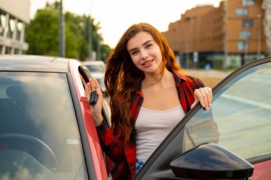 Havalı bir ekose gömlek giyen kızıl saçlı kız gösterişli kırmızı bir arabanın yanında duruyor, elleri anahtarları tutuyor.