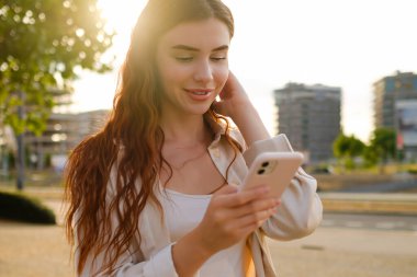 Romantik, genç, kızıl saçlı kadın gün batımında akıllı telefonunu kullanarak sokakta duruyor ve mesaj atıyor.