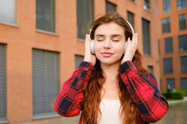 Modern kablosuz kulaklıklı, sokakta dikilmiş müzik dinleyen, kırmızı saçlı, sıradan kırmızı gömlekli, huzurlu bir kız.. 