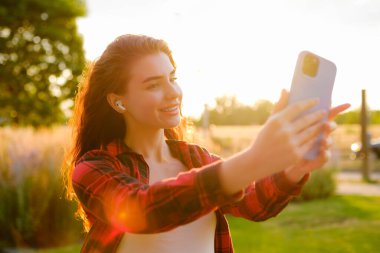 Güzel kıvırcık kızıl saçlı. Sokakta duran akıllı telefonu kullanarak selfie çekiyor.