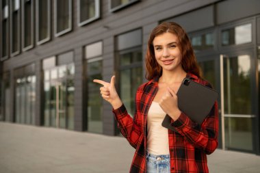 Genç bir kız öğrenci elinde tabletle kolej binasının yanında duruyor..