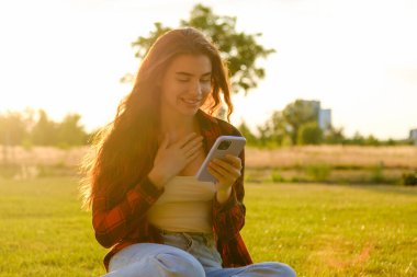 Heyecanlı genç kız, gün ışığında parkta çimlerin üzerinde oturan cep telefonuna bakar..