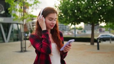 Gün batımında parkta müzik dinlemek için kablosuz kulaklık takan mutlu genç bir kadın..
