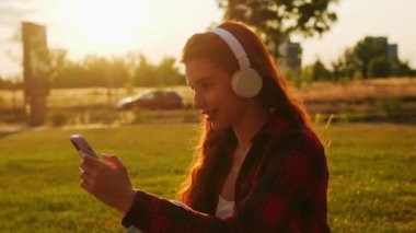 Gün batımında parkta müzik dinlemek için kablosuz kulaklık takan mutlu genç bir kadın..