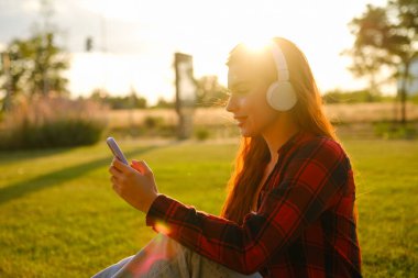 Parktaki çimlerin üzerinde oturan ve gün batımında cep telefonuyla mesajlaşan genç bir kadının profil görüntüsü. 