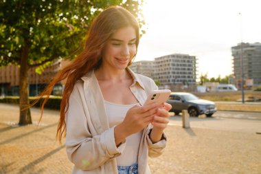 Gülümseyen kızıl saçlı kadın sokakta dikiliyor ve akıllı telefonunu kullanıyor.. 