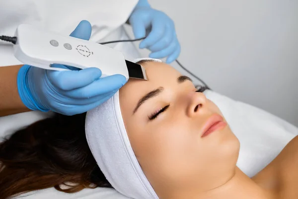 Spa worker in rubber gloves cleans face with ultrasonic device. Specialist does cleansing of body part. Smooth skin effect and blemish removal