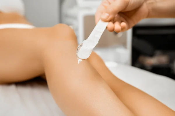 The beautician applying the cream with wax on female legs in beauty salon
