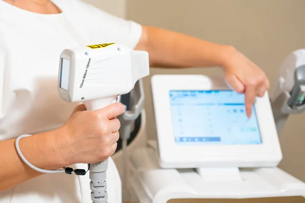 Dermatologist stands holding device from laser epilation apparatus. Cosmetologist adjusted settings on device screen for laser epilation process