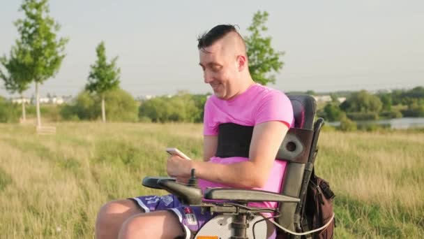 車椅子にピンクのTシャツを着ている男性は 田舎の景色に対してスマートフォンを保持しています ビデオコールで友人と話す手の障害を持つ男性 — ストック動画